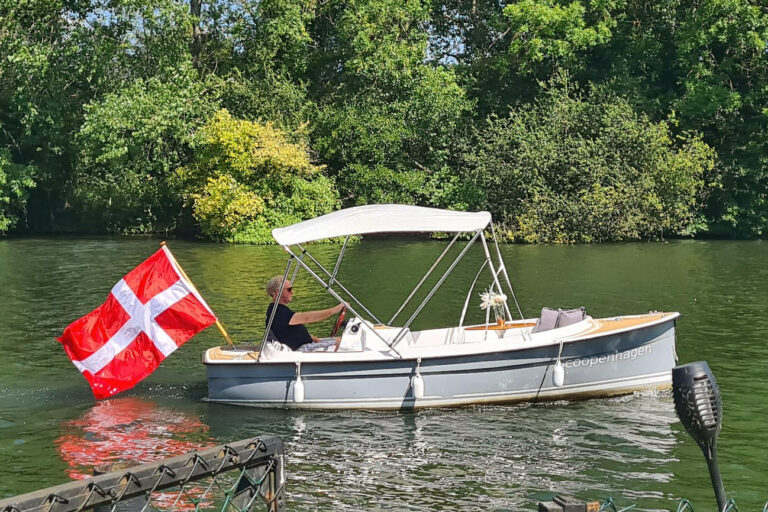 Scoopenhagen Ruban Bleu Scoop+ with Danish flag, canopy up, cruising past riverbank greenery.